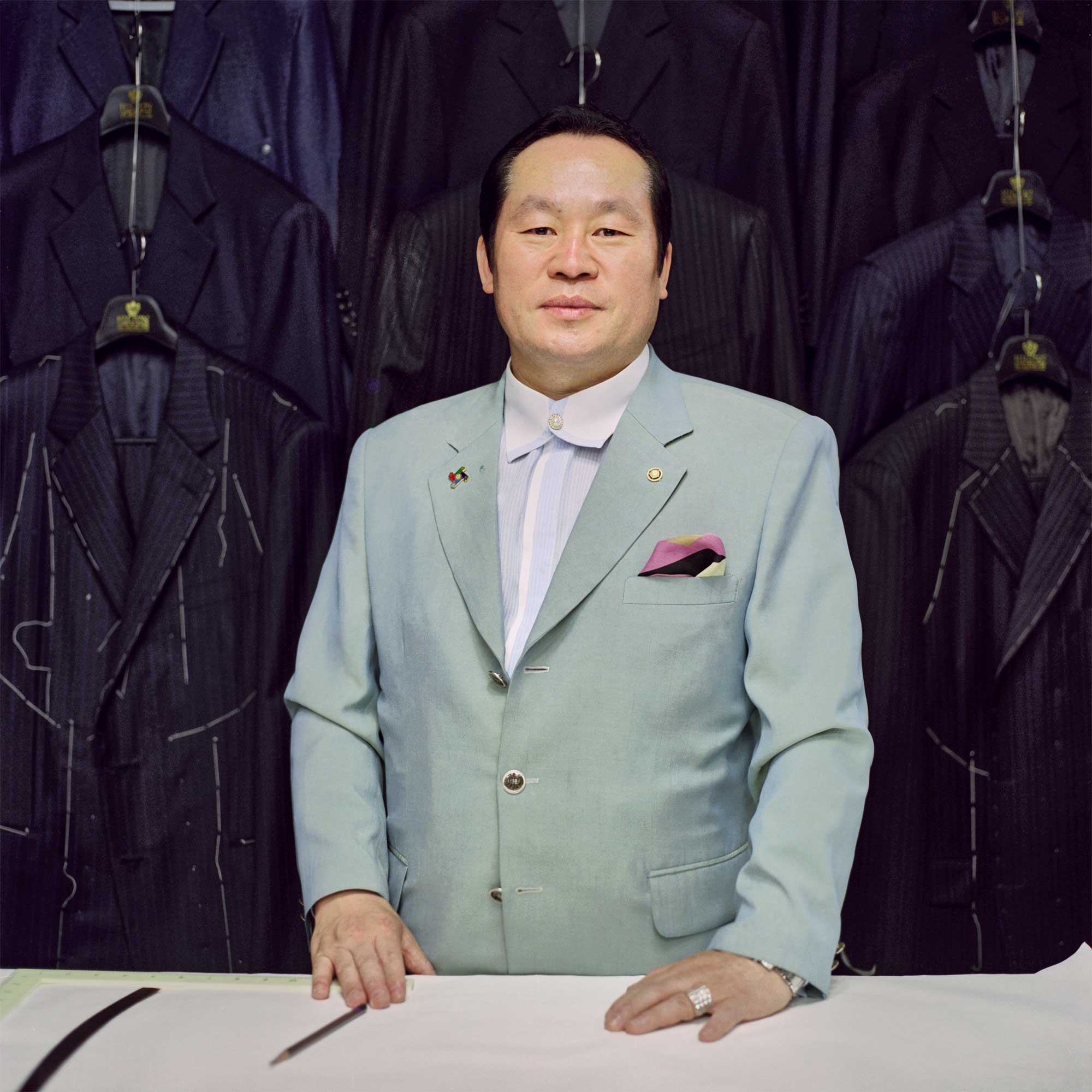 A Korean tailor in Seoul's Itaewon district stands at his tailoring table, hands resting, amidst the backdrop of suits in the making. He wears a light mint jacket and a buttoned-up shirt