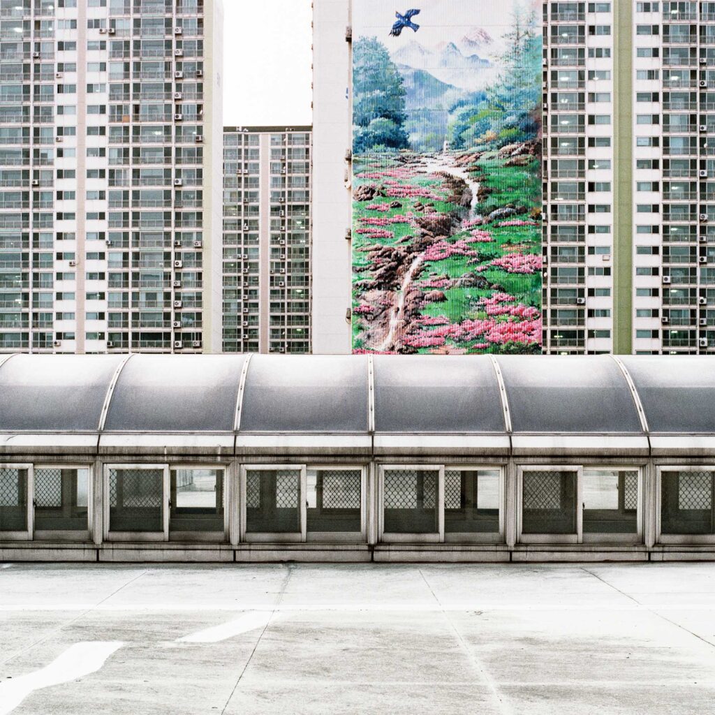 A discharge pit on a Seoul subway station rooftop, with apartment buildings rising behind. A mural on one building depicts a stream in a mountain landscape