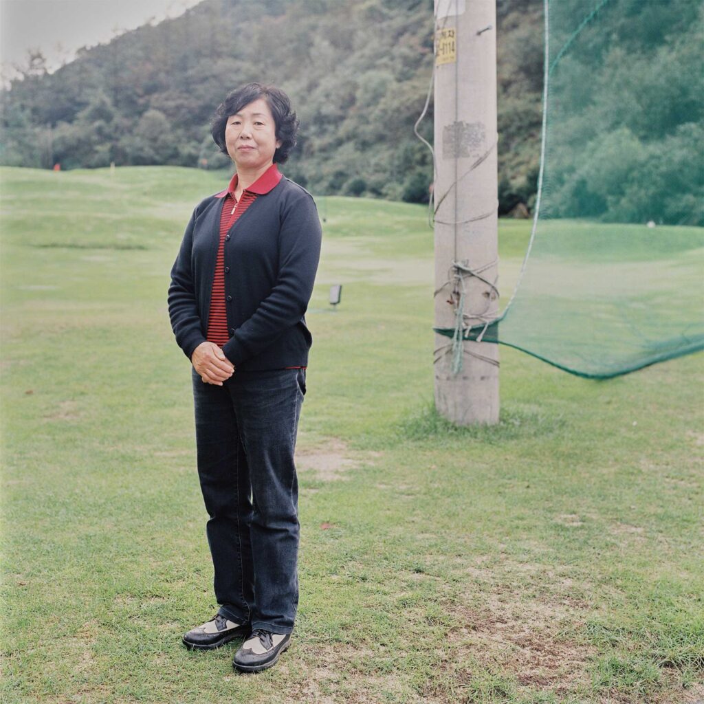 An elderly Korean Chinese woman stands beside a golf course, wearing black pants and a vest over a red and black striped polo shirt