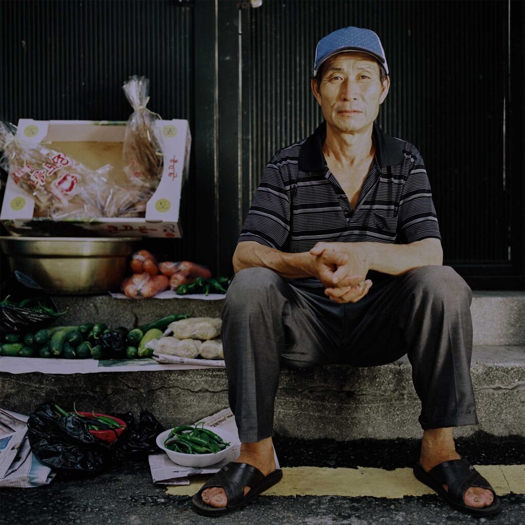 Elderly market vendor sits with vegetables on Seoul stairway, wearing baseball cap and sandals