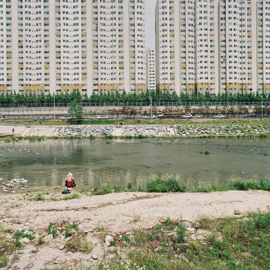 A solitary fisherman sits at the edge of a stream, its waters flowing toward the mighty Han River, framed by towering apartment buildings in the background