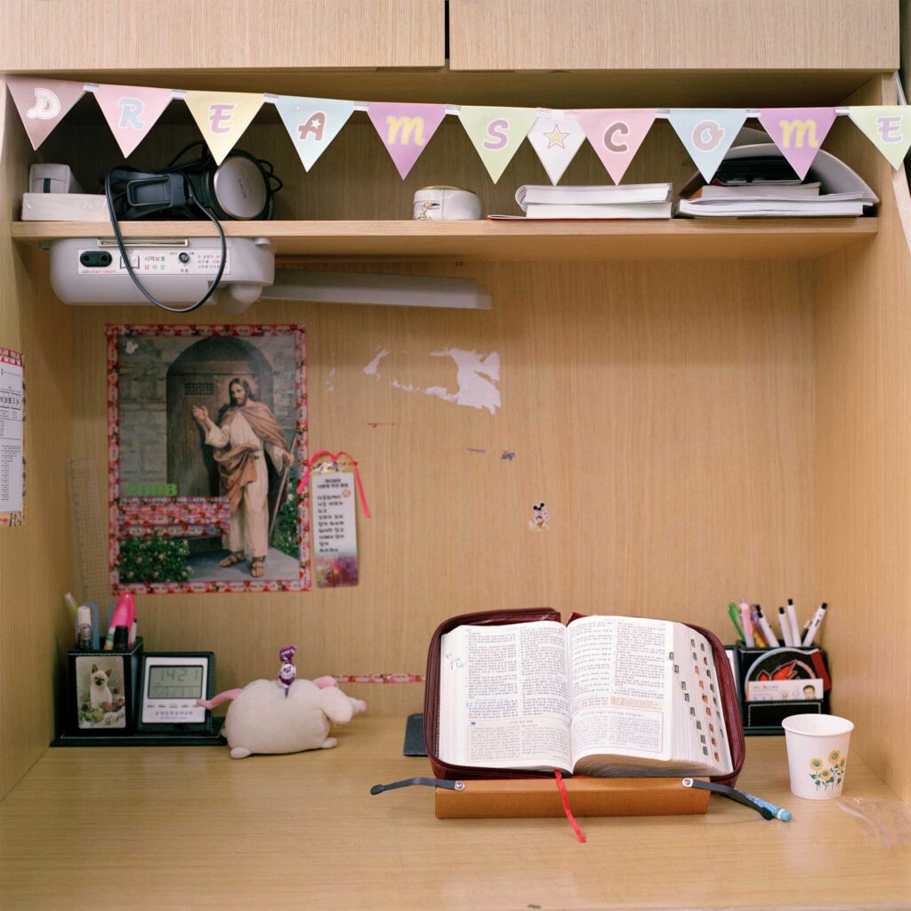 A student's desk niche featuring an open Bible, a poster of Jesus on the wall, and various pens and utensils scattered on the desk