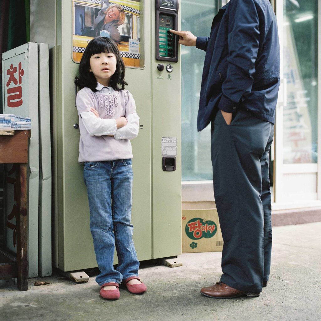 A young girl leans against a coffee vending machine outside a restaurant, arms folded. A man presses a button on the machine. Enjoying a moment outdoors