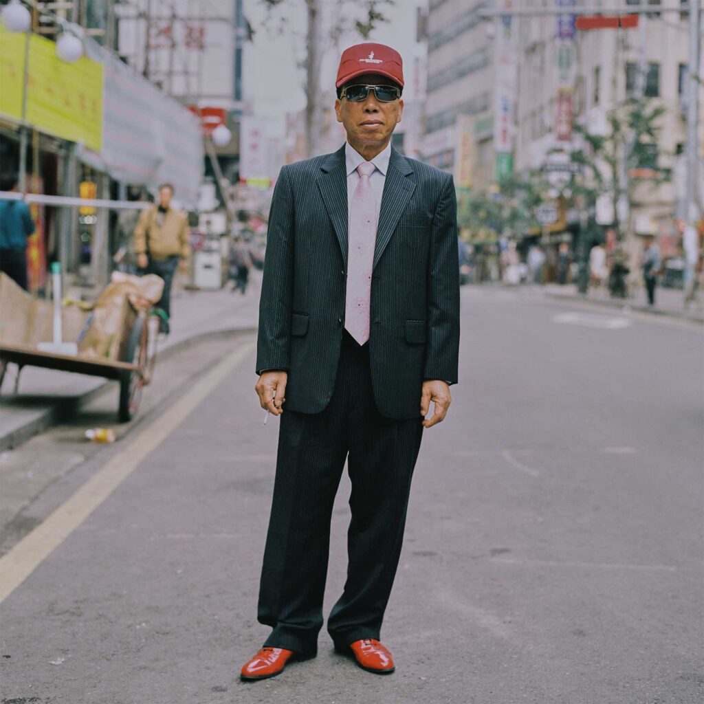 Elderly man stands in Seoul street wearing dark grey pinstripe suit, red baseball cap, and shiny red patent leather shoes