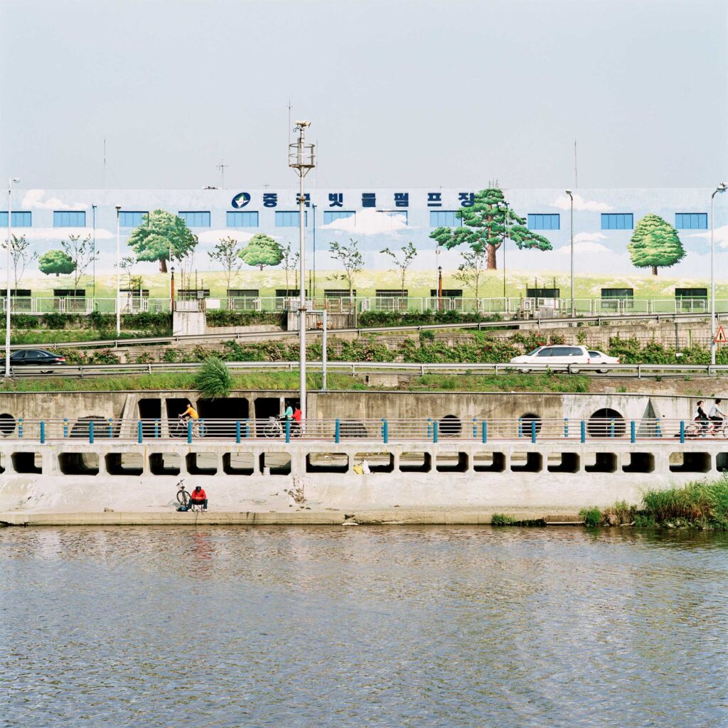 lone fisherman sits by a river in Seoul, South Korea, as cyclists pass by on the bike lane behind. In the background, a factory building displays a mural of trees on a grassy hill under a blue sky