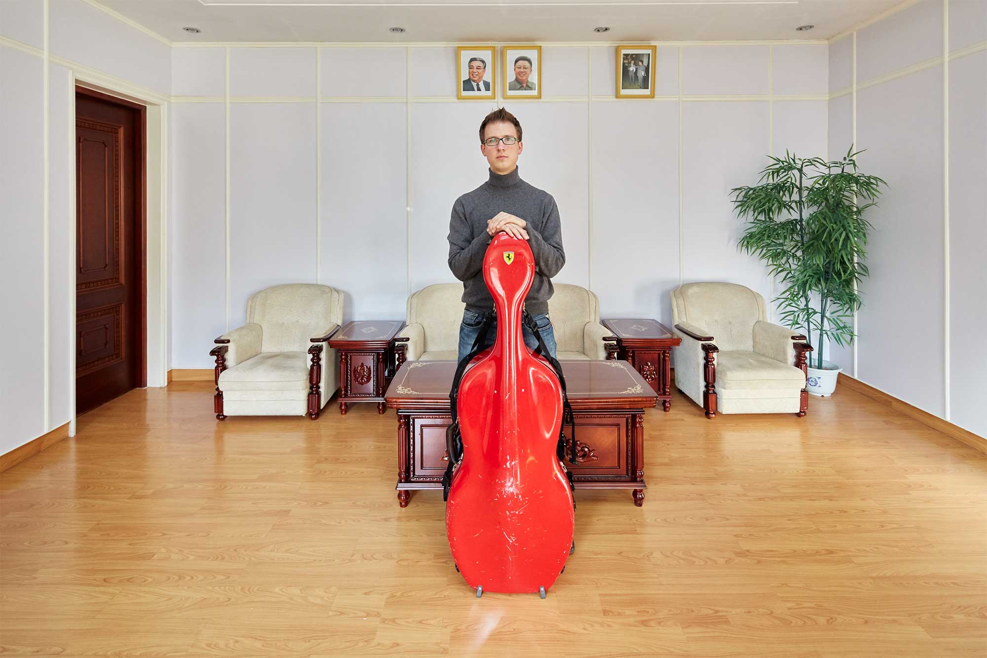 Munich Chamber Orchestra cellist in Pyongyang, North Korea, with his cello in a Ferrari-labeled red casing, surrounded by portraits of North Korean leaders Kim Il-Sung and Kim Jong-Il