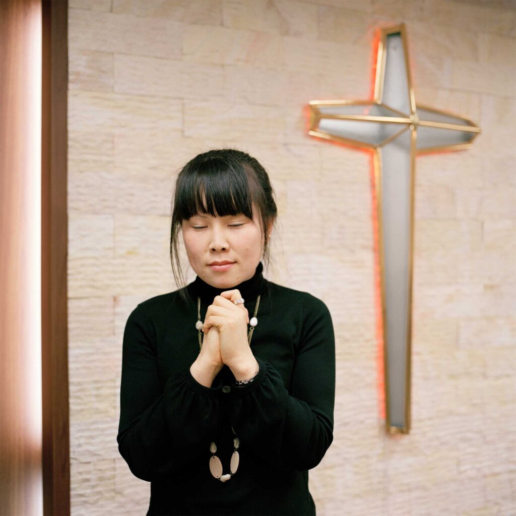 A young female North Korean defector with closed eyes, hands folded in prayer, stands before a large cross casting an orange light onto the wall behind it