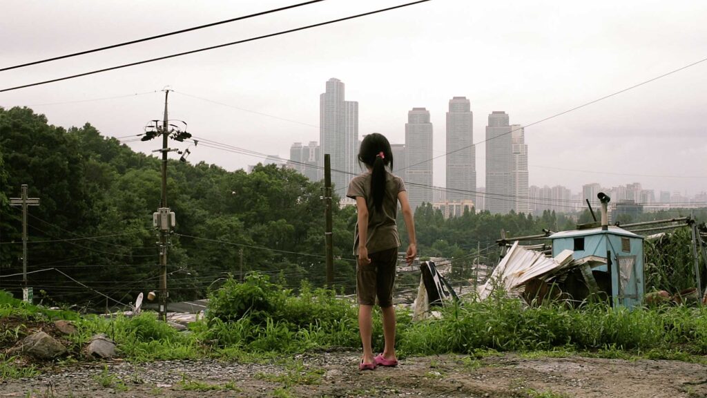 A young girl in the short film "Cold Summer" in the middle of a run-down neighborhood in Seoul, South Korea, against the backdrop of the modern high-rise skyline of Gangnam