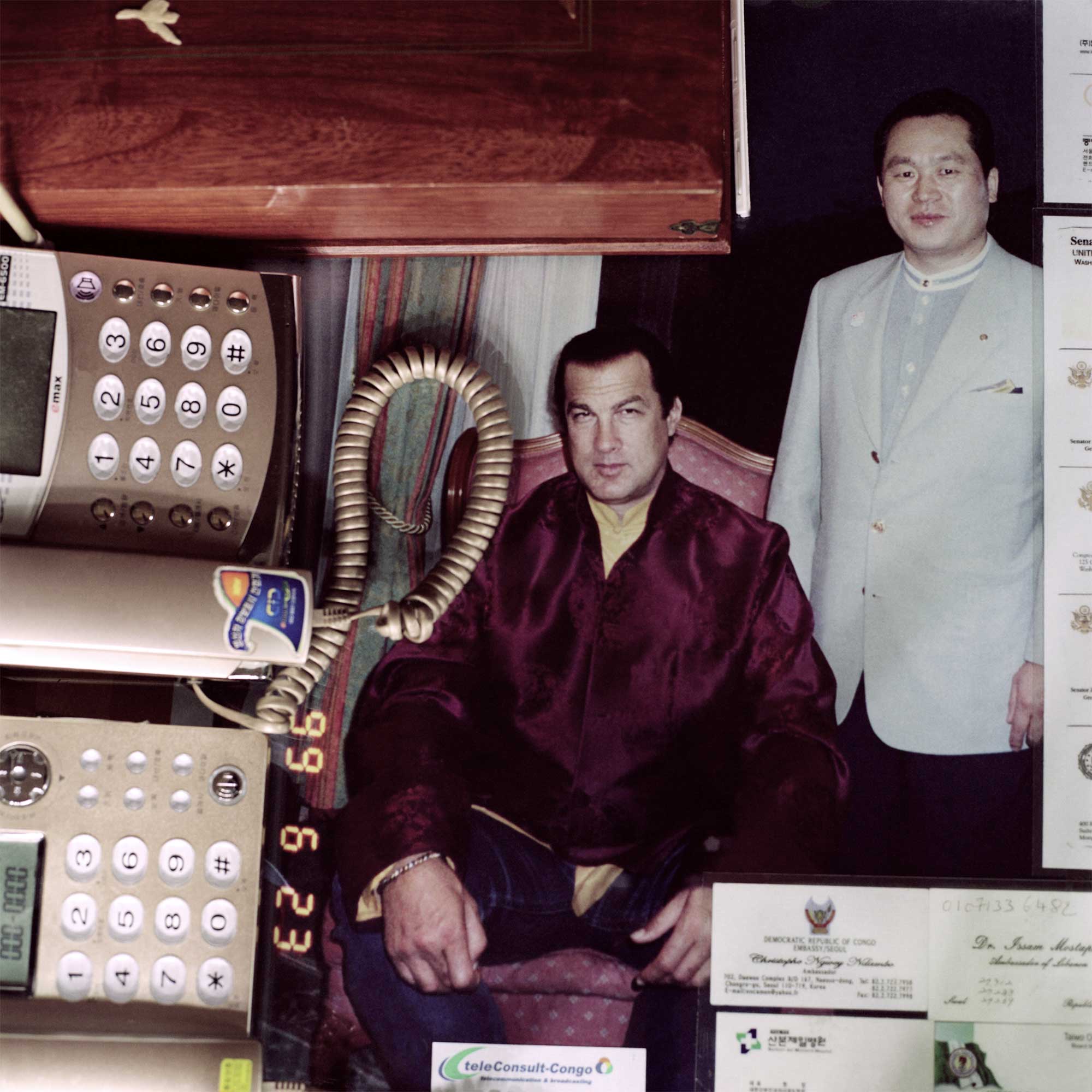 Top view of a desk with two cable telephones. Behind protective glass, a photo depicts a Korean tailor beside Steven Seagal, seated in a flamboyant purple velvet Asian-style jacket over blue jeans. Dated June 23, 1999