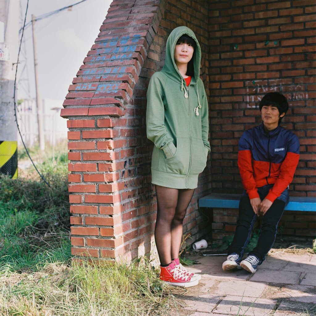 At a Korean countryside bus station, a teenage boy sits on a bench while a girl stands in front of him. She wears a fashionable hoodie over transparent black tights paired with ankle-high red Converse shoes