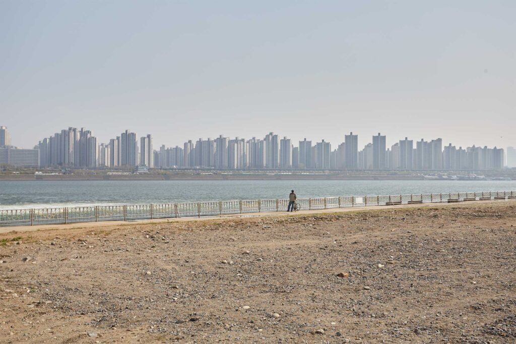 A man alone at the bank of the Han River overlooking the river and the Seoul apartment complex buildings rising up on the opposite side