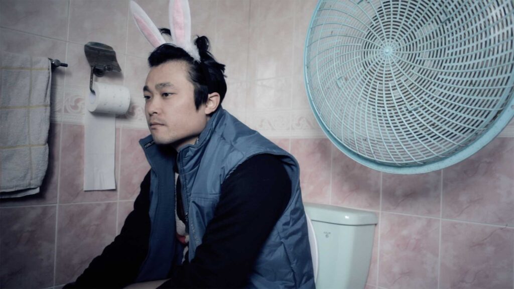 A scene from the Wolf + Lamb vs. Soul Clap 'Lonely C' music video: The brother, wearing a bunny hair ribbon, sits lost in thought on a toilet in the bathroom of a remote South Korean countryside village