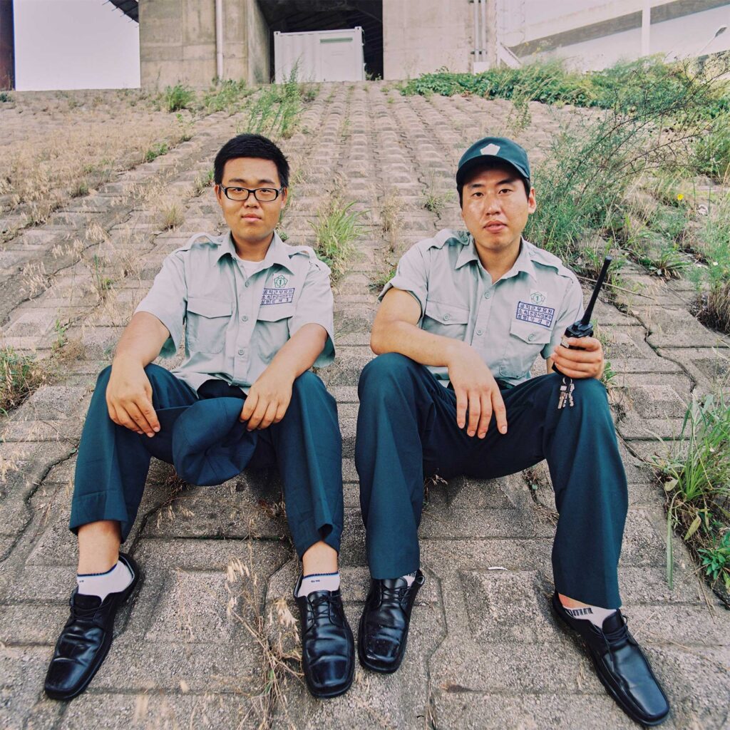 Two male public service workers in identical uniforms sit beneath a flyover road in Seoul, South Korea, crossing the Han River. One holds a hat, the other a walkie-talkie