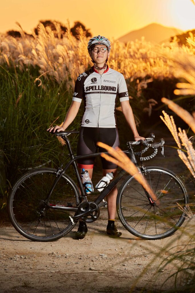 A professional Korean cyclist stands behind his bicycle in a field of reeds as the sun sets, coloring the sky.