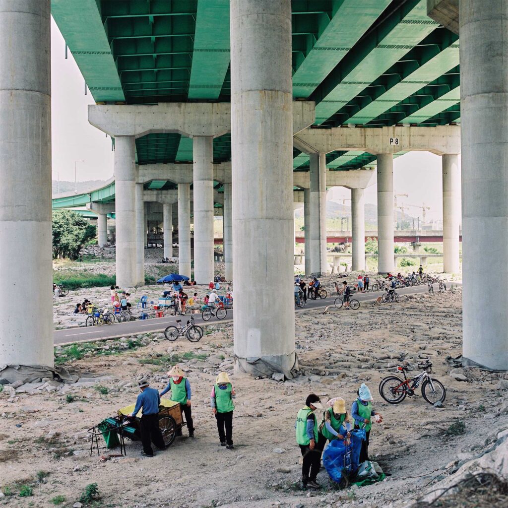 A highway bridge over a stream flowing to the Han River. Bikers find shade on a hot day as workers clean up trash