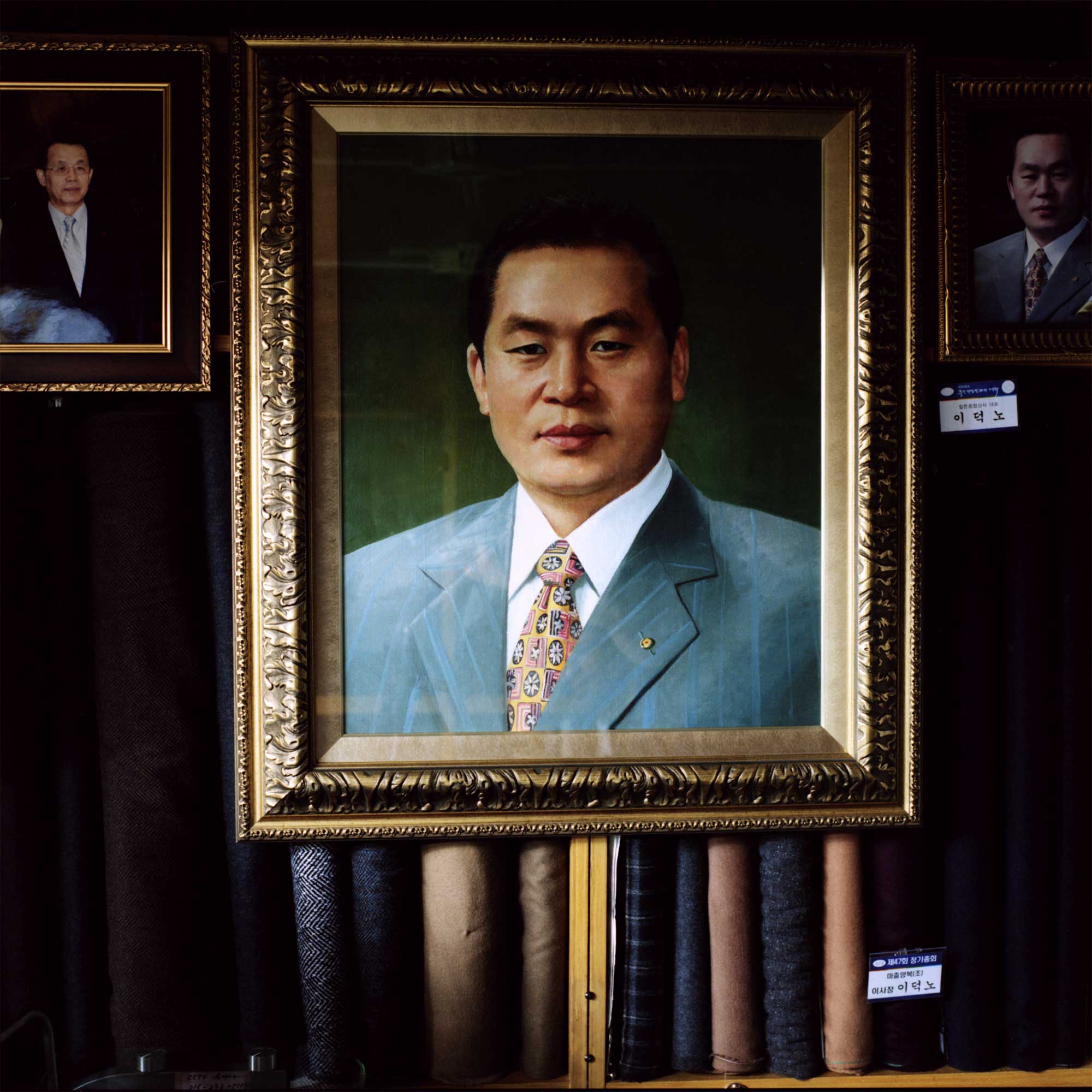 A framed acrylic portrait of a Korean tailor hangs on a shelf adorned with colorful fabrics. Adjacent, a smaller painting repeats the image. To the left, another framed image features a Korean personality