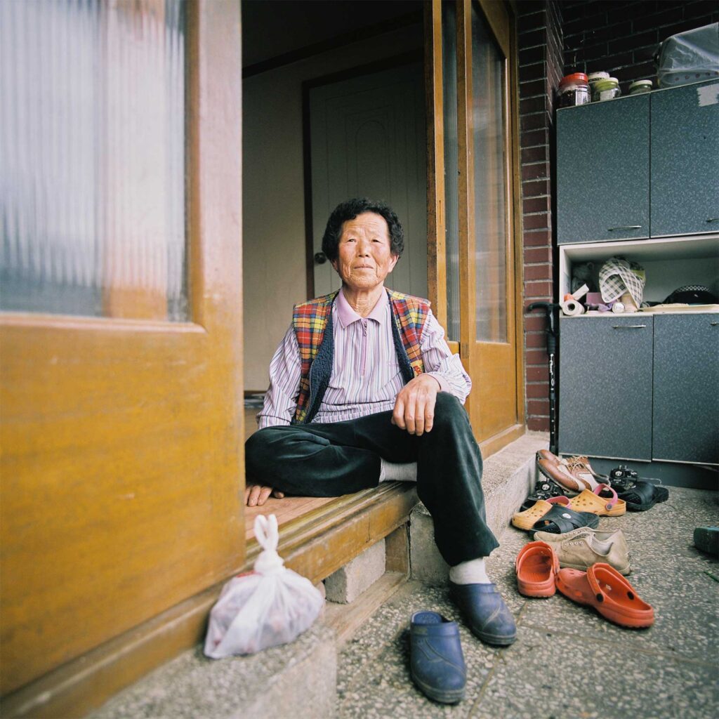 In the Korean countryside, an elderly woman sits on the floor between wooden and glass sliding doors at the entrance to her house. Pairs of shoes are piled up right at the entrance
