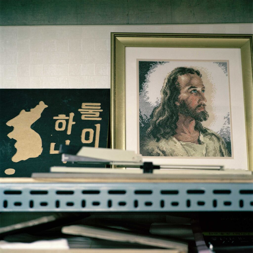 An insert shot of a shelf displaying a woodcarving depicting the North Korean Peninsula without division between North and South. Adjacent to it is a picture frame containing a stitching of Jesus Christ