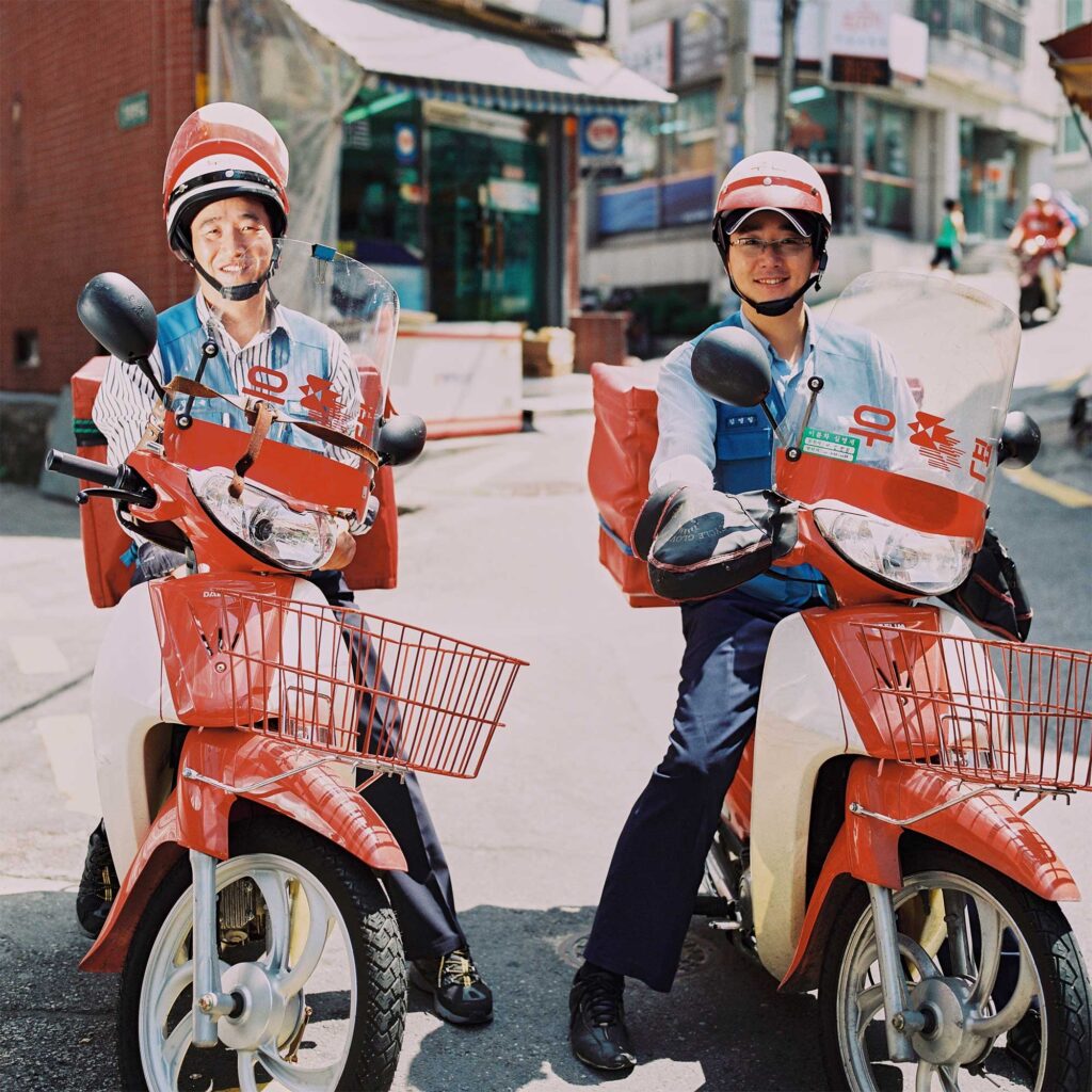 Two Korean postal service delivery drivers, uniformed, sit on their resting motorcycles in a side street in Seoul, South Korea. Helmet visors flipped up
