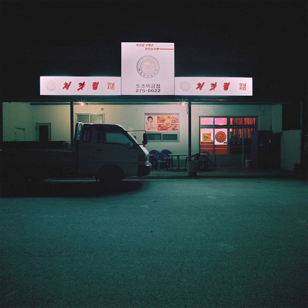 At night in the Korean countryside, a brightly lit restaurant with a billboard on the roof, contrasting against the darkness. A pick-up truck is parked in front