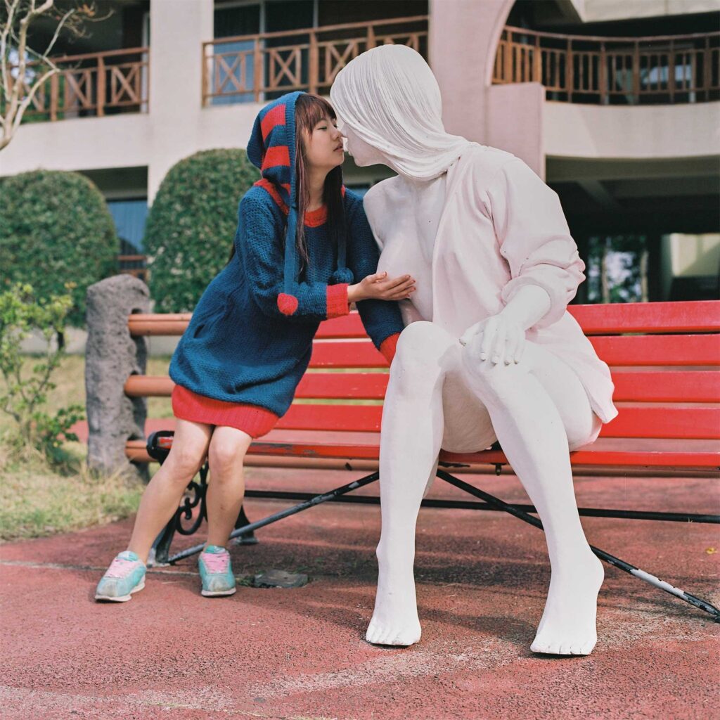 A girl sits next to a female sculpture on a bench and gives the sculpture a kiss at Loveland sculpture park, Jejudo Island, South Korea.