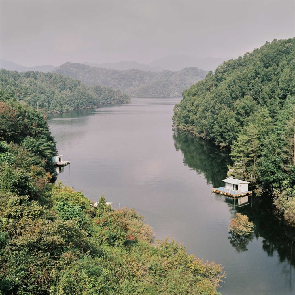 In the Korean countryside, an artificial dam creates a serene lake. Dense forests cover the hilly landscape, reaching to the water's edge. Two floating fishing platforms are anchored near the lake's shore