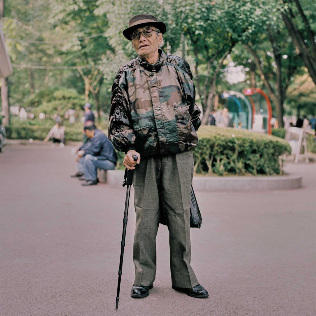 Old Korean man in Seoul park, leaning on walking stick, wears camouflage bomber jacket, hat, olive green pants, holds black plastic bag