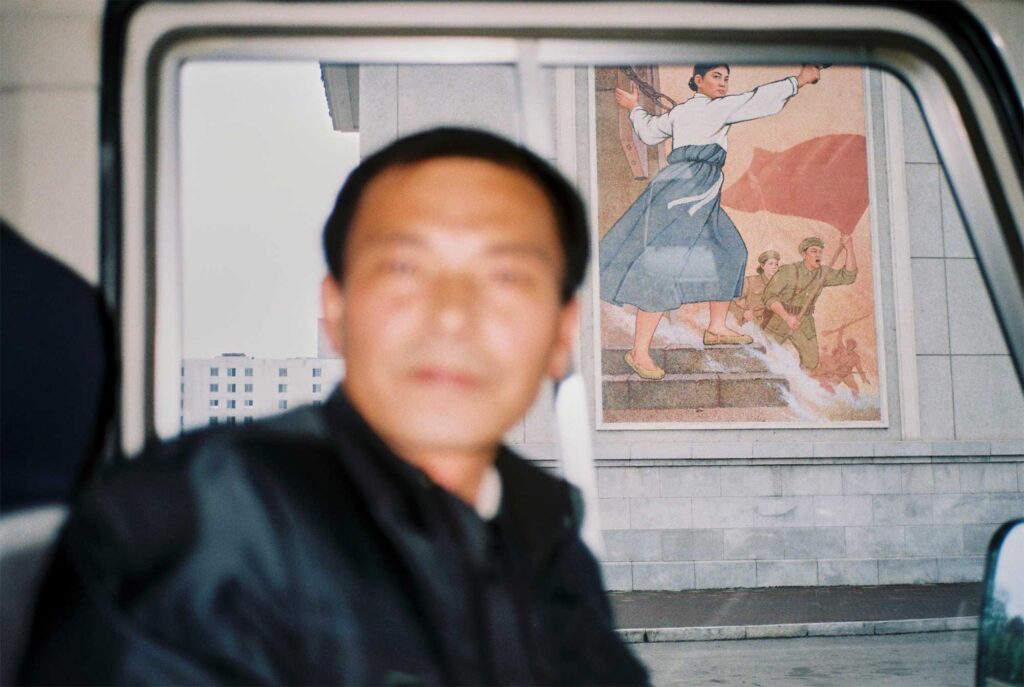Tour-guide bus parked in front of Pyongyang's National Library, showcasing a propaganda mural in the background