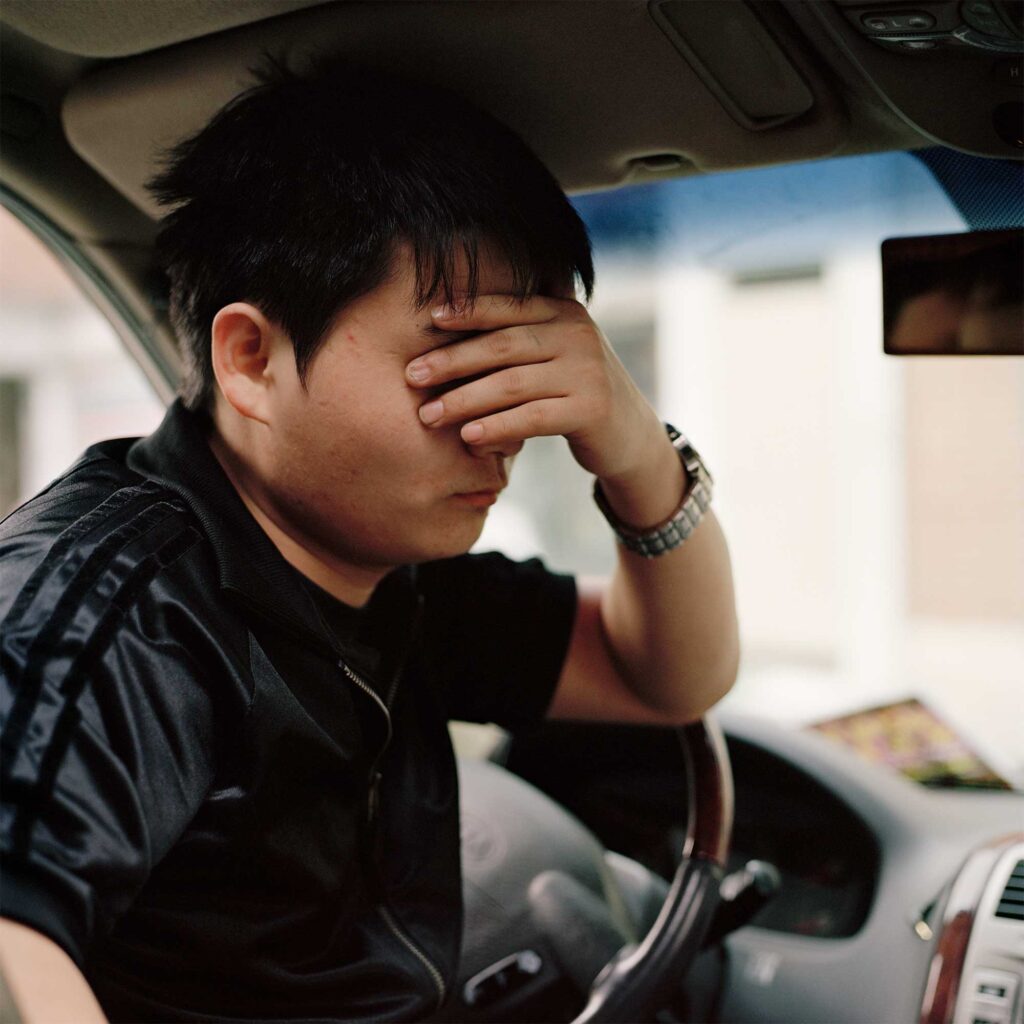A young North Korean male defector sits in the driver's seat of a parked car, covering his face with his left hand