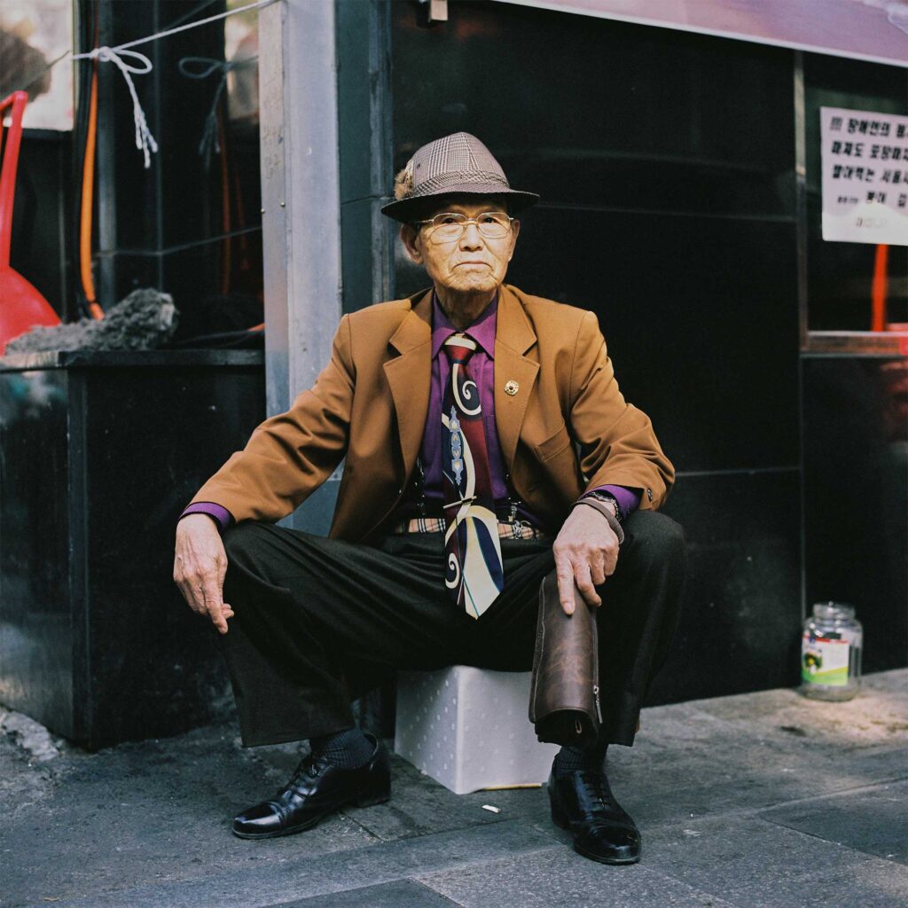 Elderly Korean man sits on stone at Seoul street corner. Wearing checkered hat, brown suit jacket, purple shirt, patterned necktie, checkered belt, dark pants. Sunlight on face
