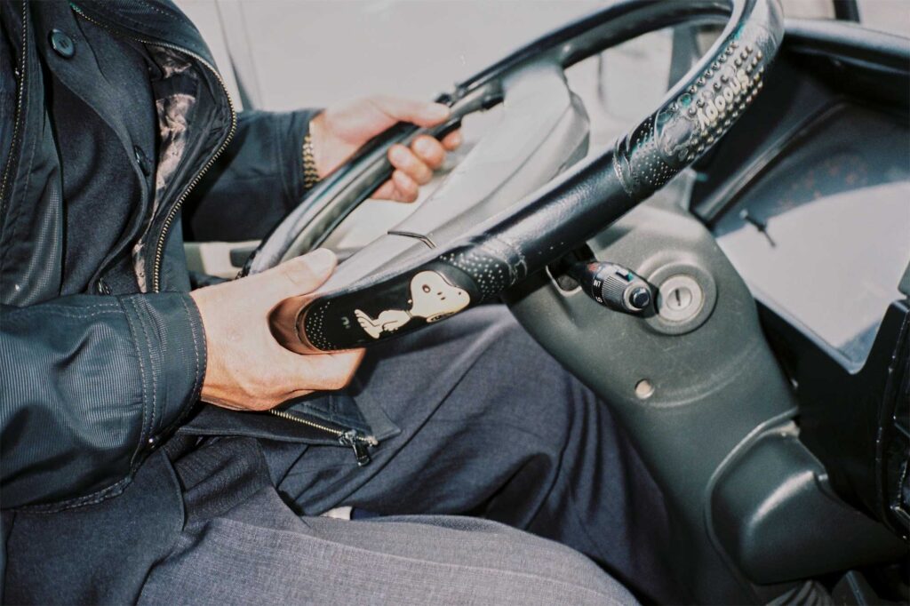 Hands on the steering wheel of a tour-guide bus parked in front of Pyongyang's National Library, adorned with a Snoopy character sticker, in North Korea.