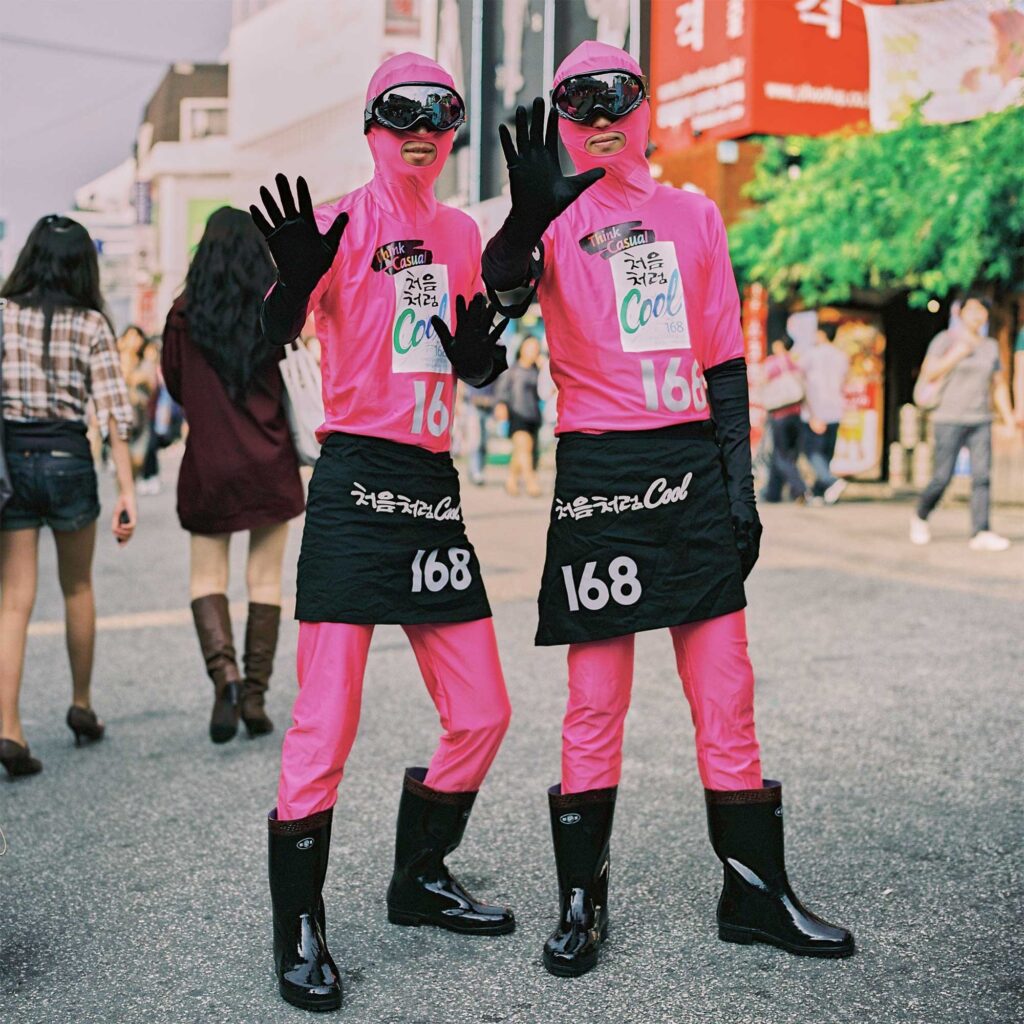 Two males stroll the streets of Seoul in funky, space-themed uniforms to advertise a product. Clad in pink pants, tight space hoodies, black dresses, gum boots, and ski visors, they grab attention