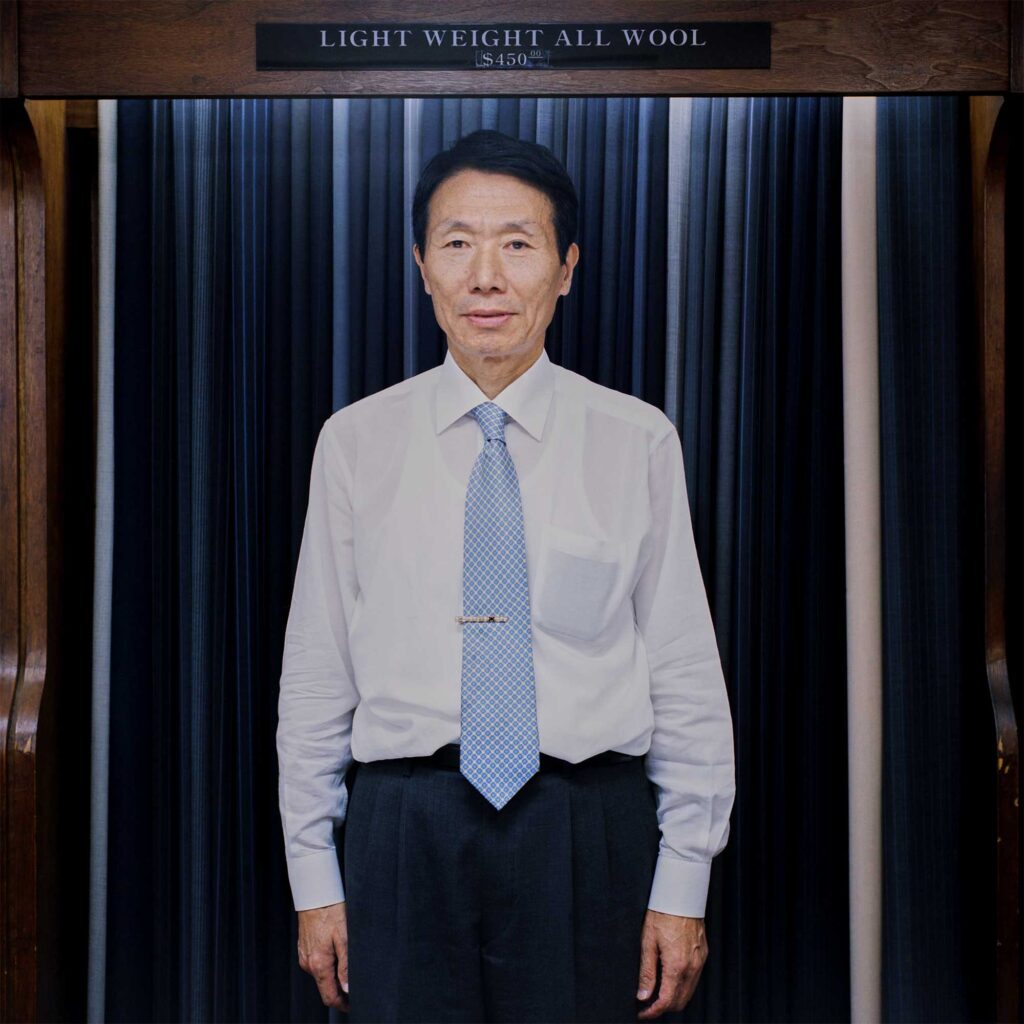 A Korean tailor stands before an open wardrobe labeled 'Lightweight All Wool $450'. Dressed in a white shirt, black pants, and a white and blue patterned necktie, he exudes professional elegance
