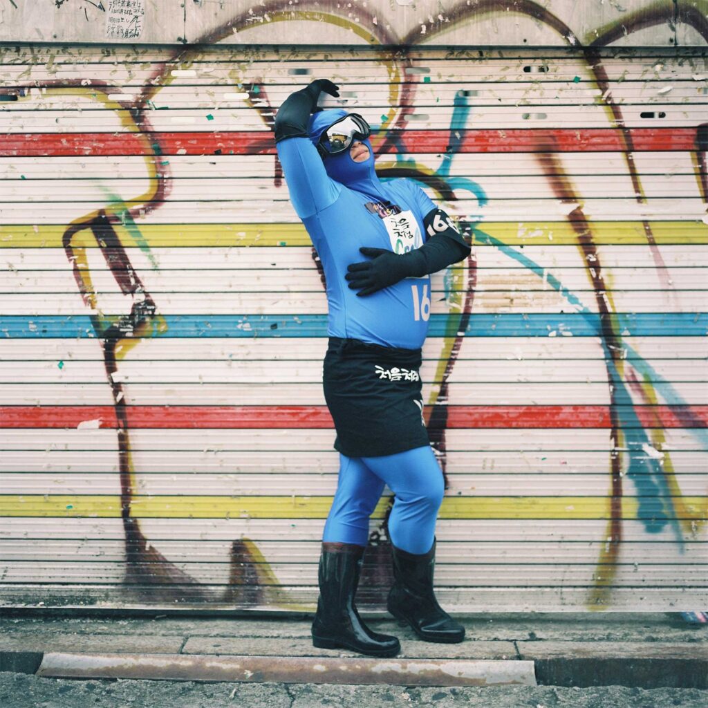 A man dressed in a space-like costume poses in front of the camera in front of a colorful striped garage in the Hongdae district of Seoul, South Korea
