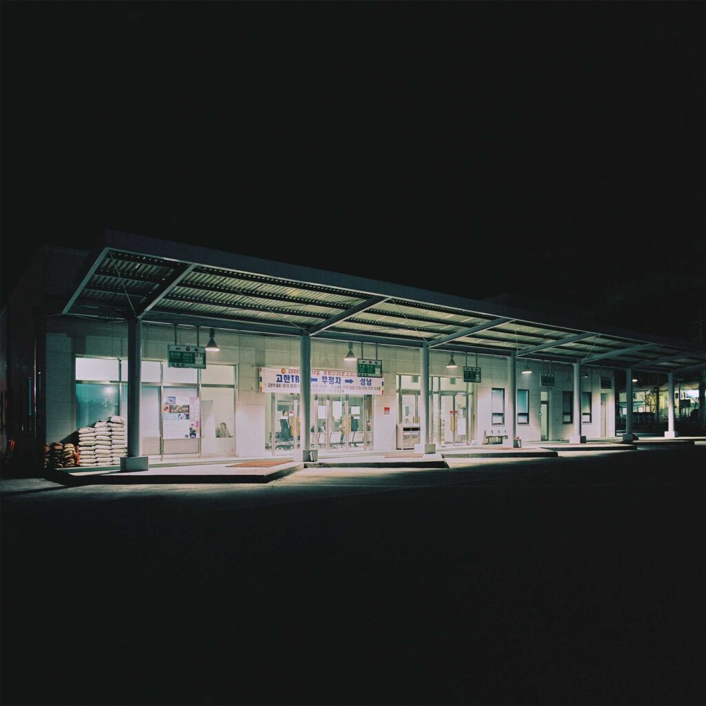 In the dark of the night, an empty bus terminal in the Korean countryside stands illuminated, its lights highlighting the architectural features of the building.