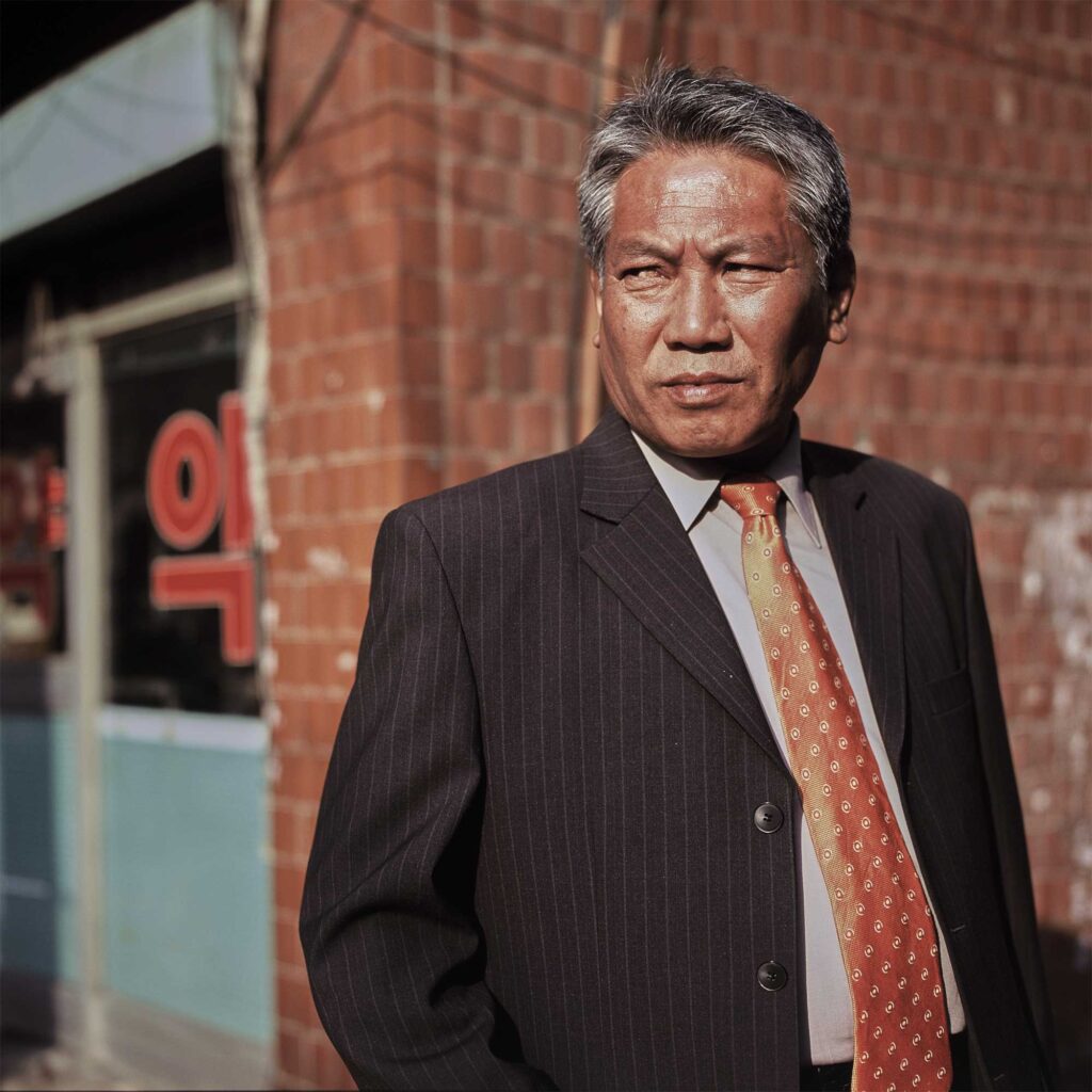 Man in front of Seoul pharmacy. Grey pinstripe suit, light grey shirt, terra-cotta necktie. Healthy grey hair, tan face