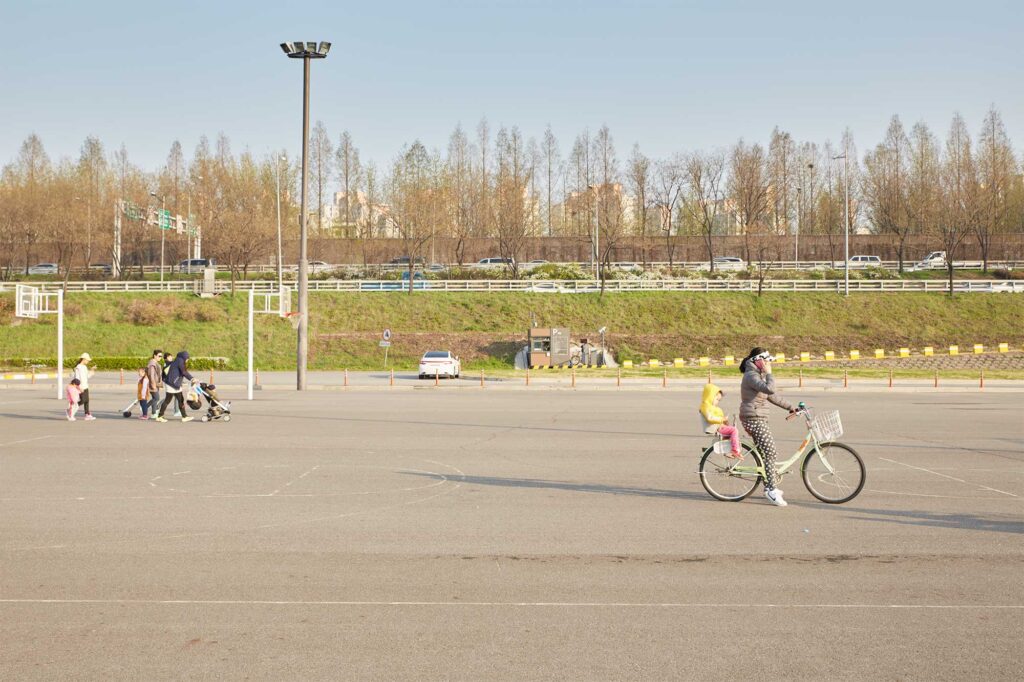 A mom on a call resting on her bike with her kid on the back seat in a paved sports ground next to Han River in Seoul, South Korea
