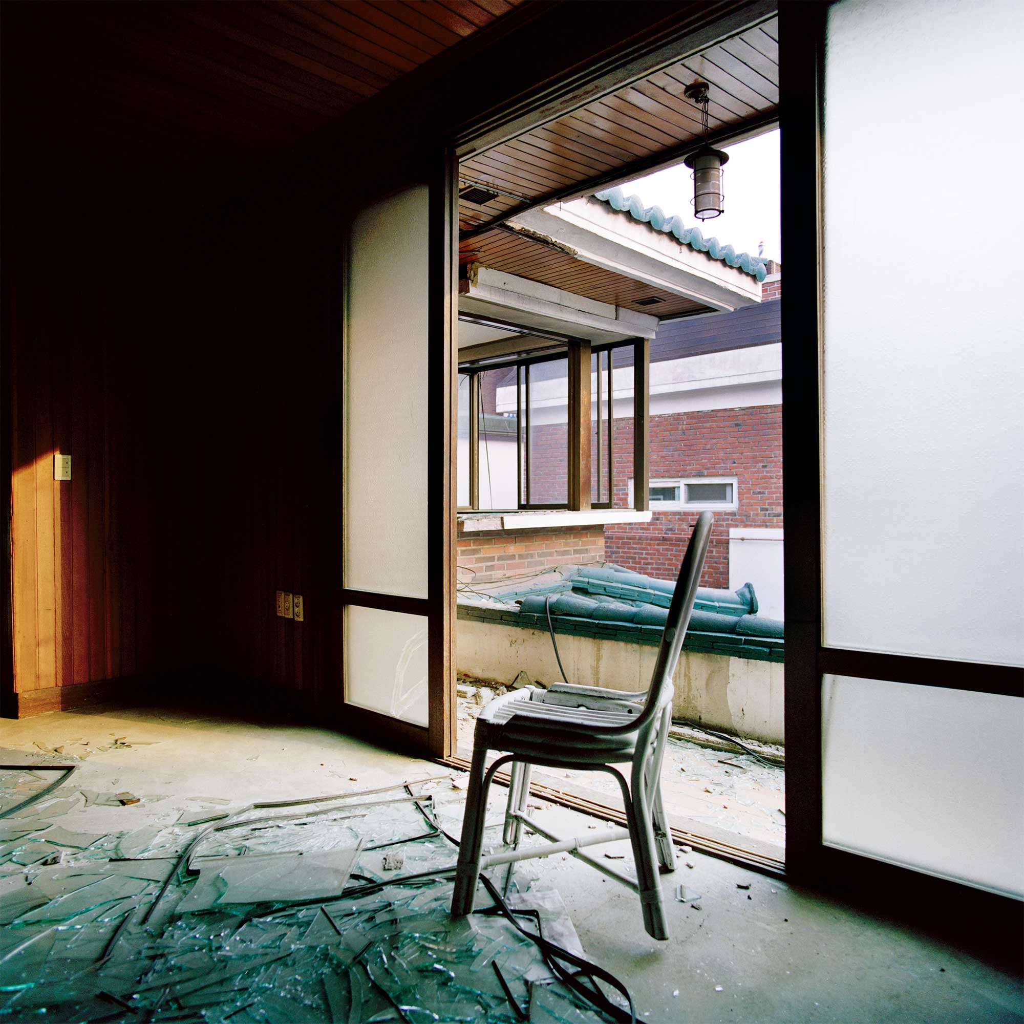 In a Seoul apartment undergoing redevelopment, the abandoned living room speaks of neglect. Broken glass litters the floor, with an empty chair amidst. Sunlight filters through the wooden panels, offering warmth in contrast to the cold