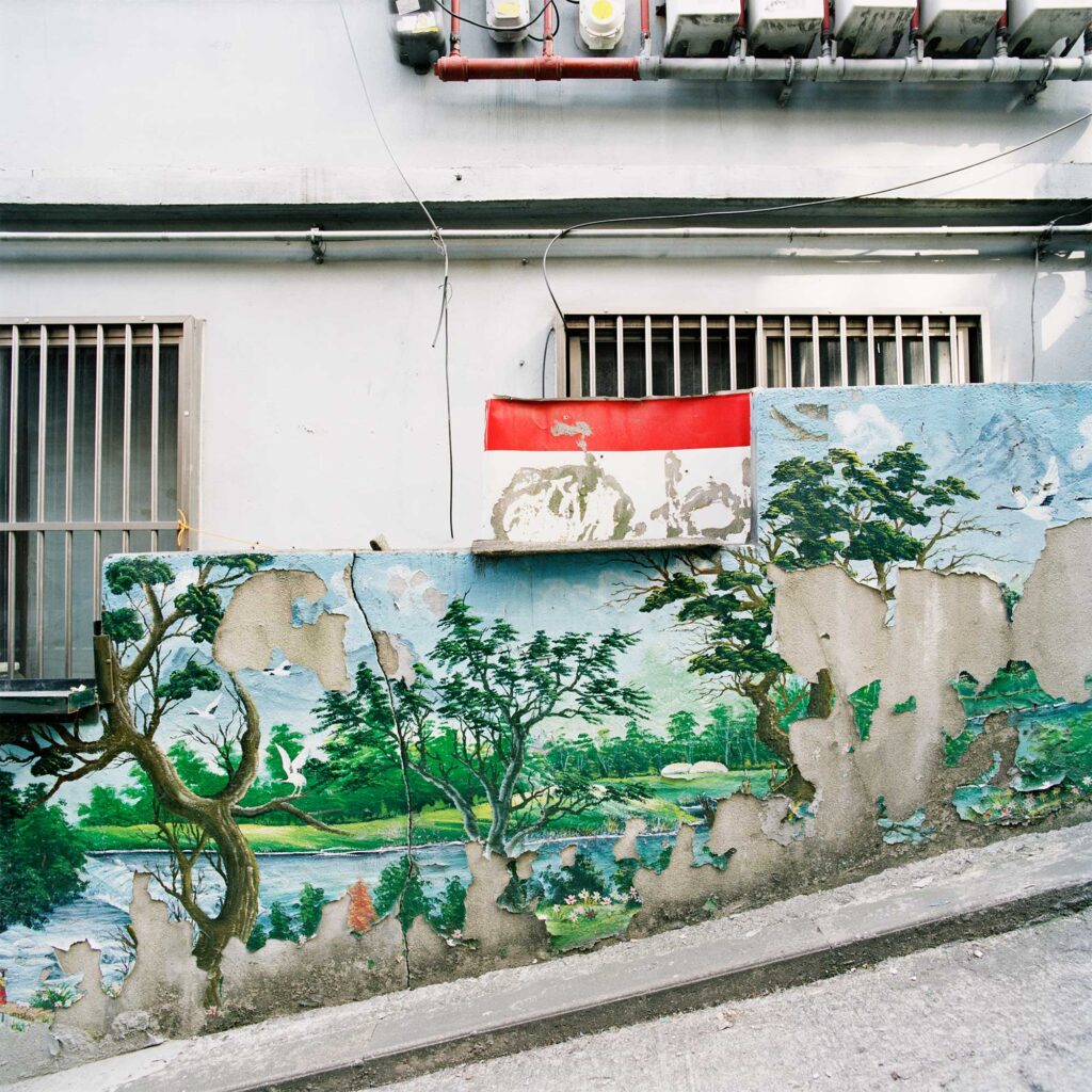 The facade of an older residential building on a steep decline in Seoul, South Korea. A mural on the surrounding wall depicts a serene landscape with crumbling paint