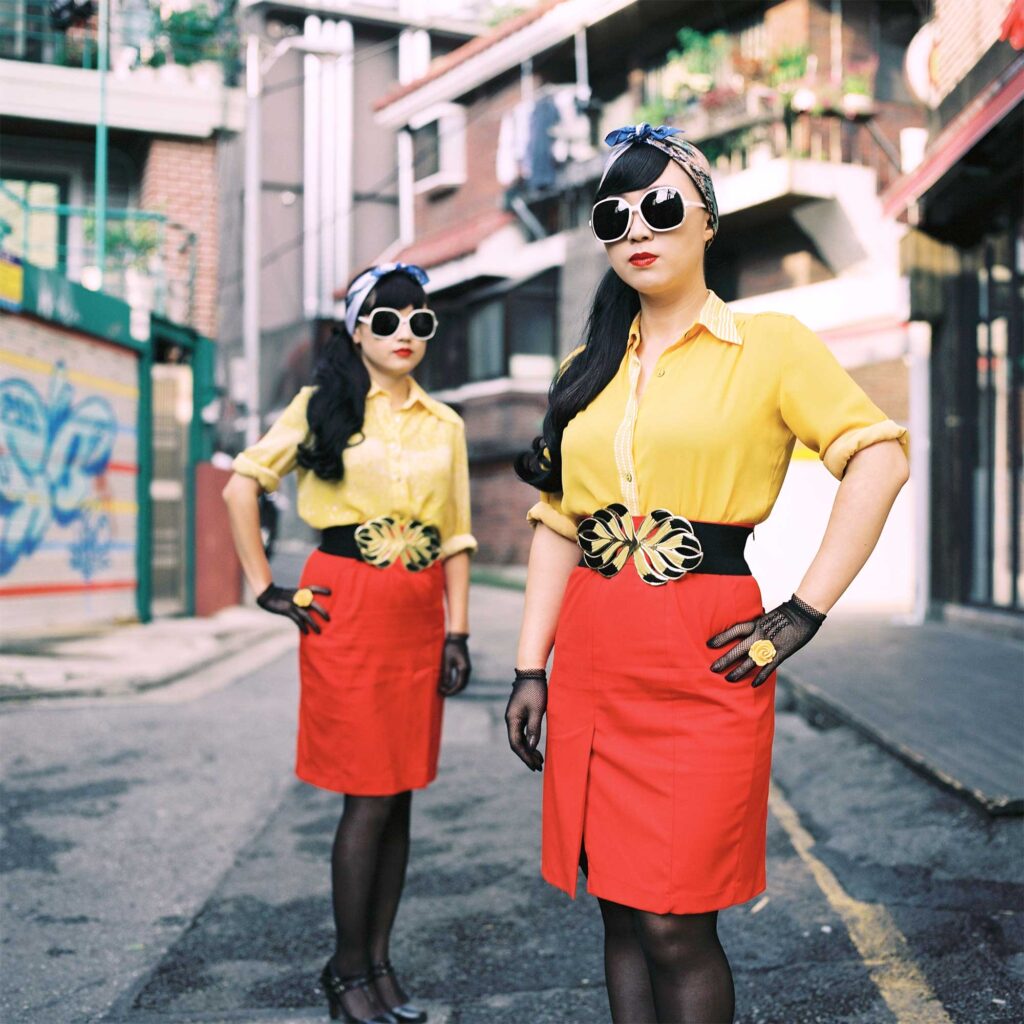 Two young women pose in a Seoul back alley. Vintage vibes: red skirts, yellow blouses, feather-like belt buckles, sunglasses, hairbands, gloves with yellow flowers. Hands on waist.