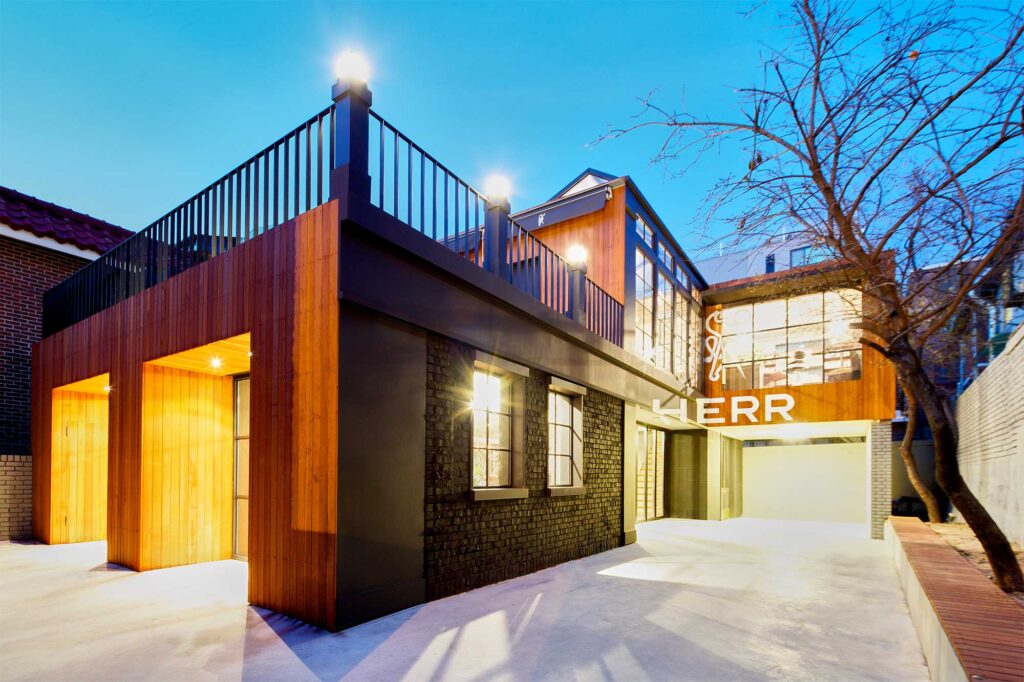 Early evening exterior view of the gentlemen's barber shop HERR in Seoul, South Korea, showcasing a facade of wood and brick with the store logo prominently highlighting the entrance area.