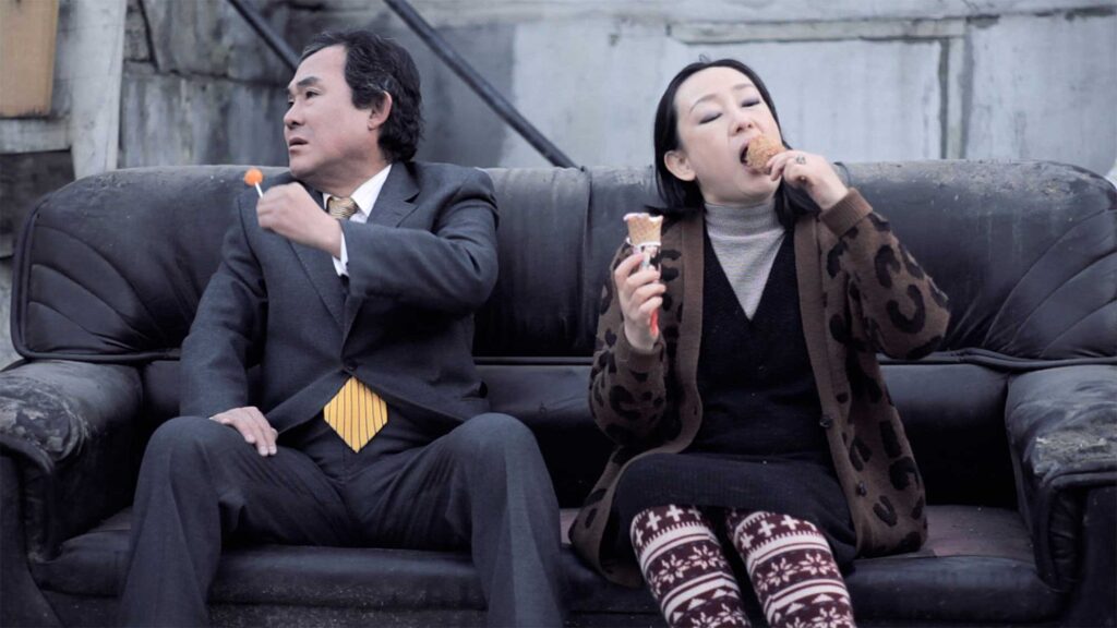 A scene from Röyksopp's 'Senior Living' music video: The older man sits next to the older woman as she indulges in an ice-cream and a hotdog on a stick, satisfying her pregnancy cravings. The man, annoyed by her attitude, pops a lollipop into his mouth.
