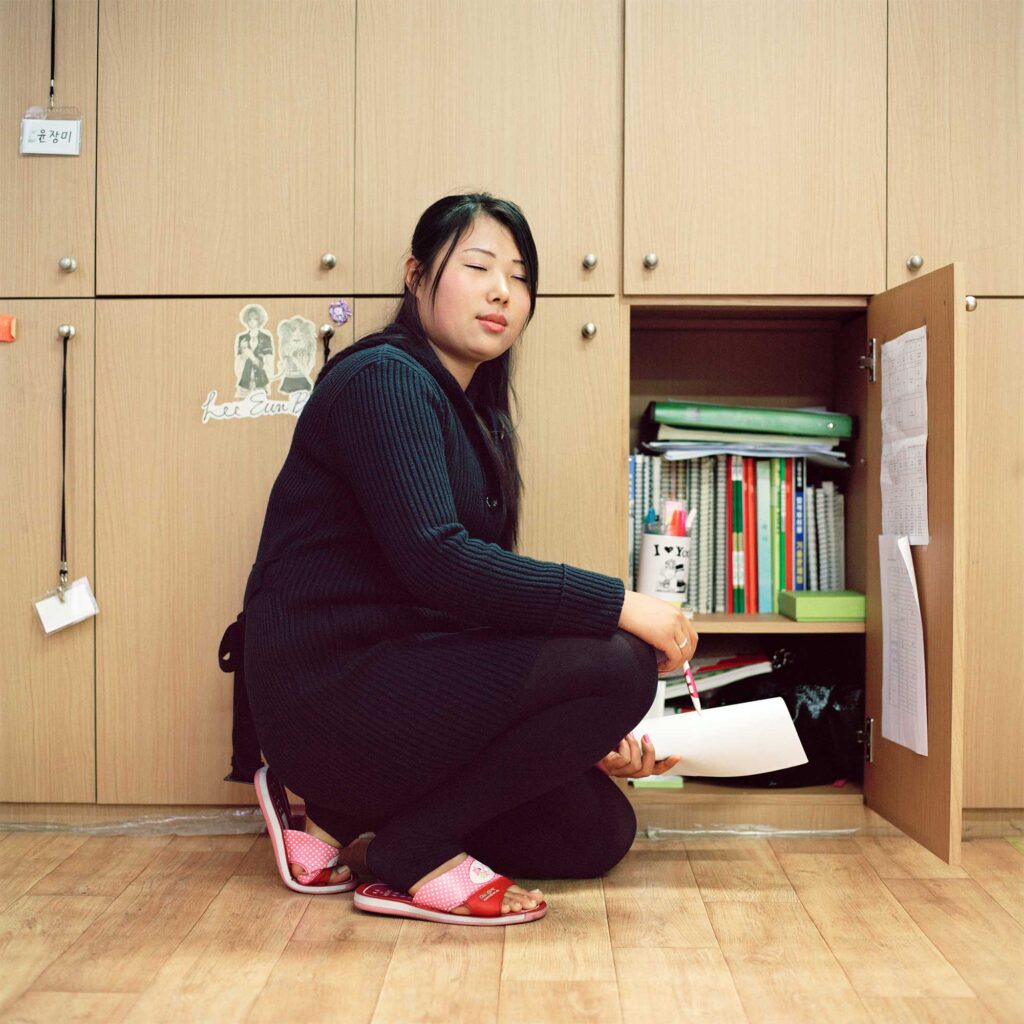 A North Korean female defector student kneels in front of her open locker in school, facing the camera with her eyes closed