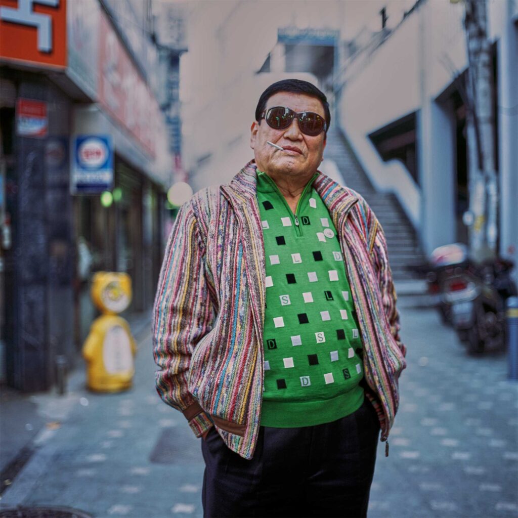 A cool man with a green checkered sweater and a stripped jacket wearing sunglasses and having a cigarette sticking out of his mouth