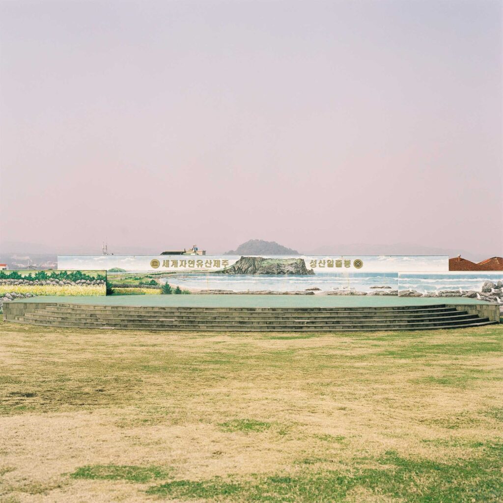A grassy field with a performance stage backed by a wall depicting Seongsan Ilchulbong on Jejudo Island. A real hill rises above the painted landscape