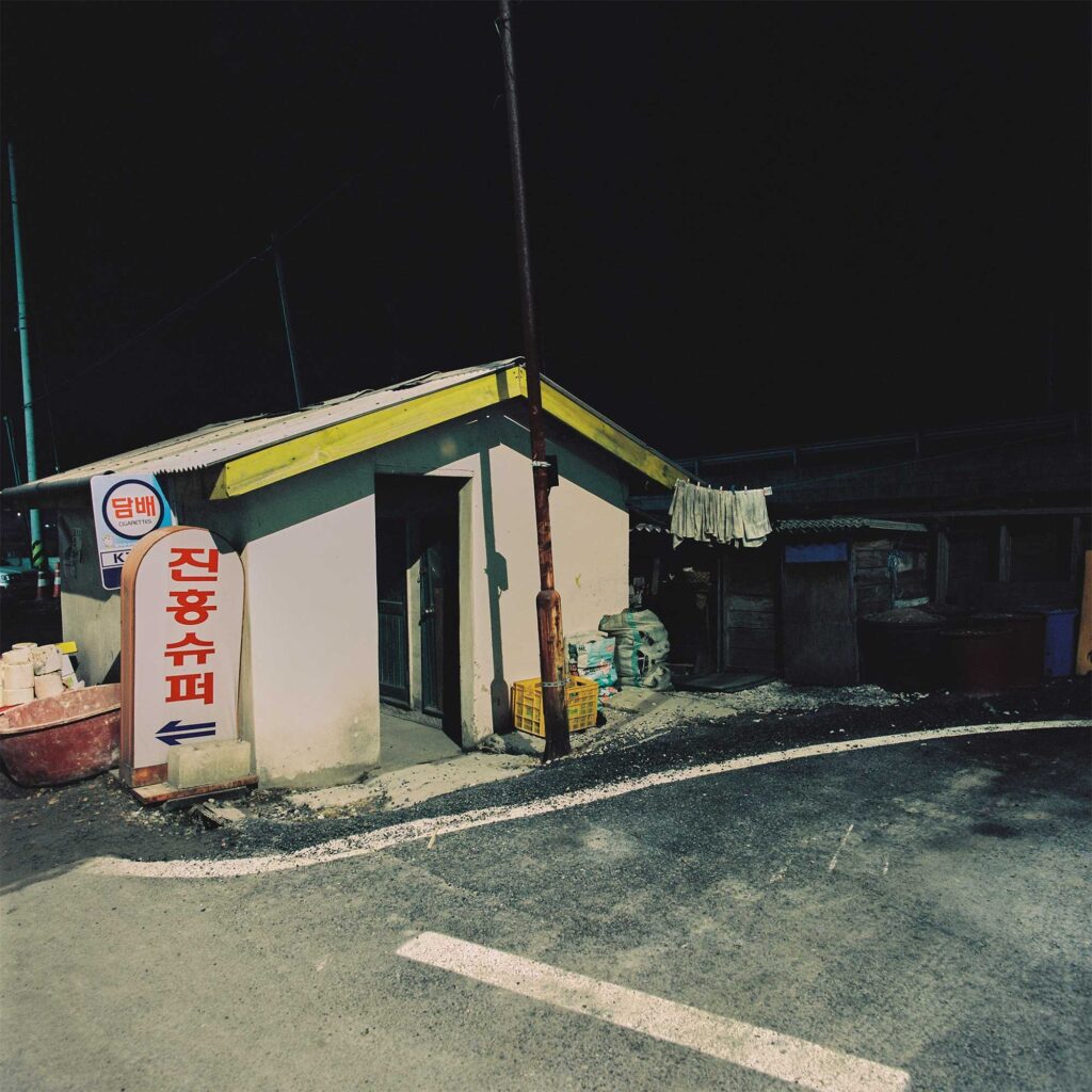 At night, a small corner convenience store in the Korean countryside stands out, illuminated by the glow of a nearby street lantern