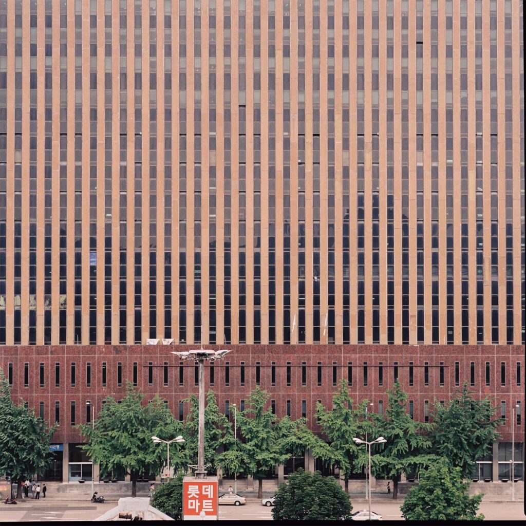 Brown-toned facade rising above a line of trees, with windows punctuating the concrete in a linear pattern