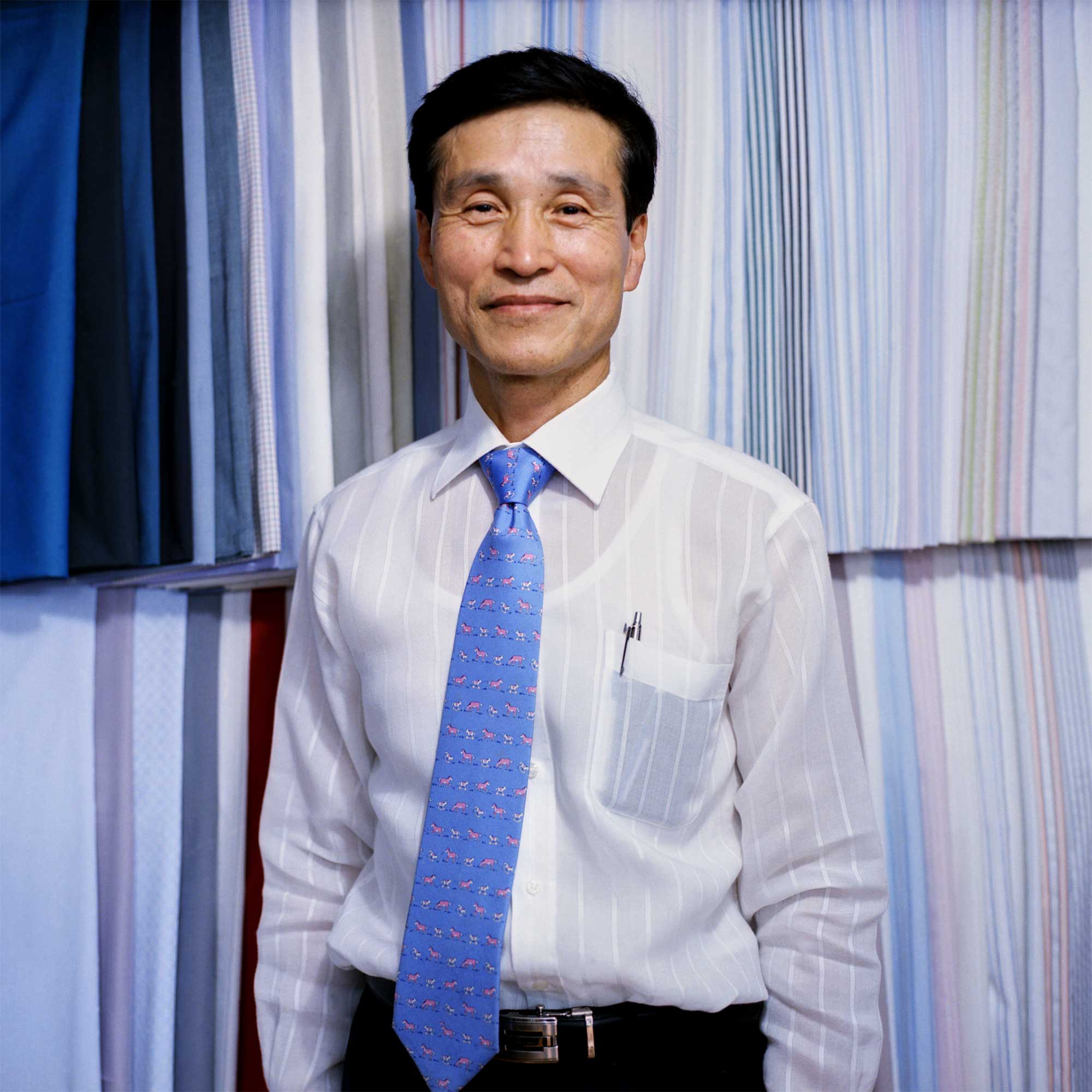 A Korean tailor stands amidst an array of colorful fabrics, his white dress shirt slightly transparent, revealing his undershirt. A pen peeks from his shirt pocket, while a blue necktie with a horse pattern adds a touch of personality