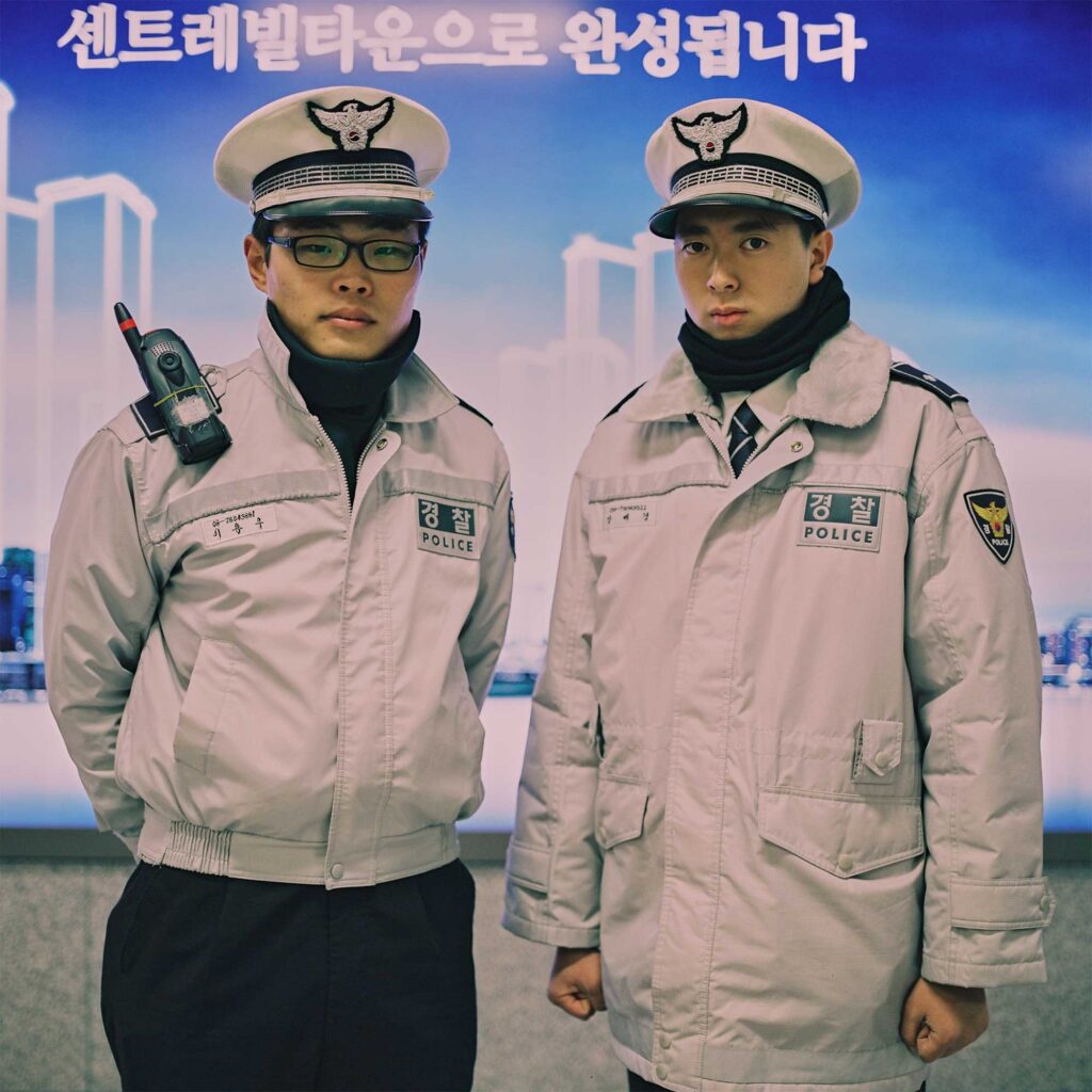 Two male police officers stand in front of an advertising billboard in a Seoul subway station. Dressed in warm winter uniforms, one wears a shorter jacket while the other opts for a longer padded one