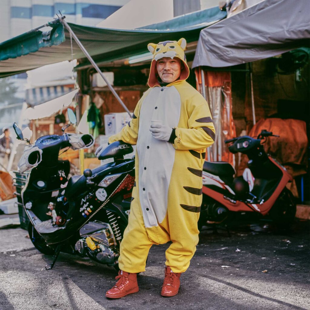 A middle-aged Korean man in a tiger costume and red sneakers pauses for a vending machine coffee break beside his motorcycle